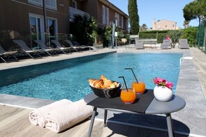 Escena junto a la piscina con sillones de descanso, una mesa con cruasanes, zumo de naranja y un jarrón con flores.