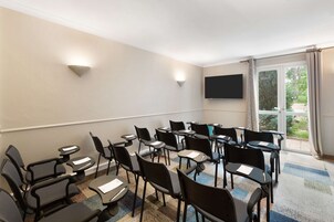 Un salón de conferencias con varias mesas y sillas, un televisor de pantalla plana montado en la pared y un gran ventanal con cortinas.