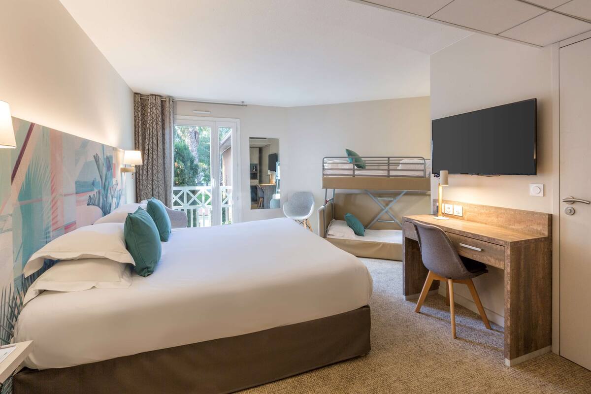A hotel room with a large bed, a desk with a chair, a television, and a balcony with a view of greenery.