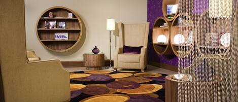 A modern hotel room with a round wooden bookshelf, a beige armchair, a purple patterned wall, and a decorative beaded structure.