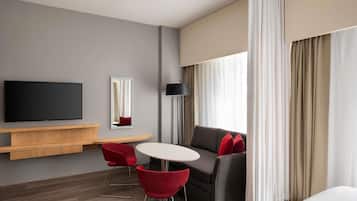 A hotel room with a flat-screen TV, a sofa, a dining table, red chairs, a lamp, and a large window with curtains.