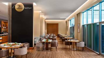 A modern hotel lobby with a reception desk, a seating area with round tables and chairs, and large windows offering views.