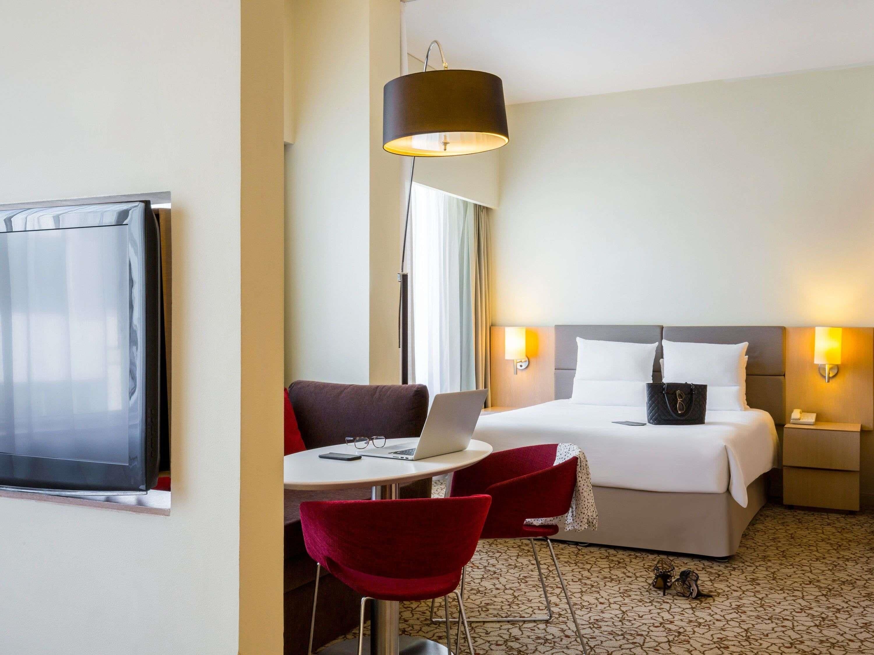 A hotel room with a bed, bedside tables, a desk with a laptop, a chair, and a TV.