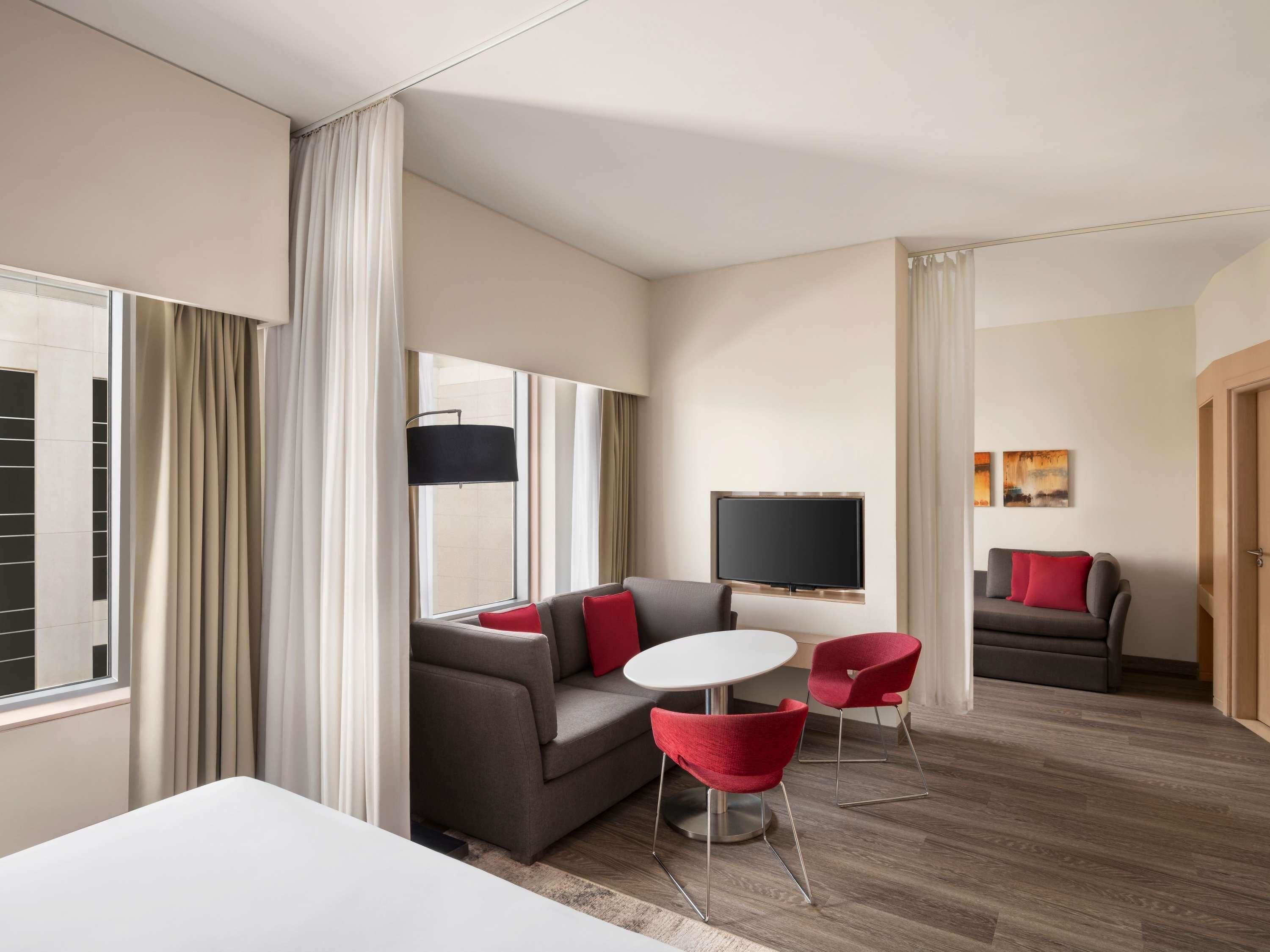 Une chambre d’hôtel avec un canapé, deux fauteuils rouges, une petite table, un téléviseur et une porte.