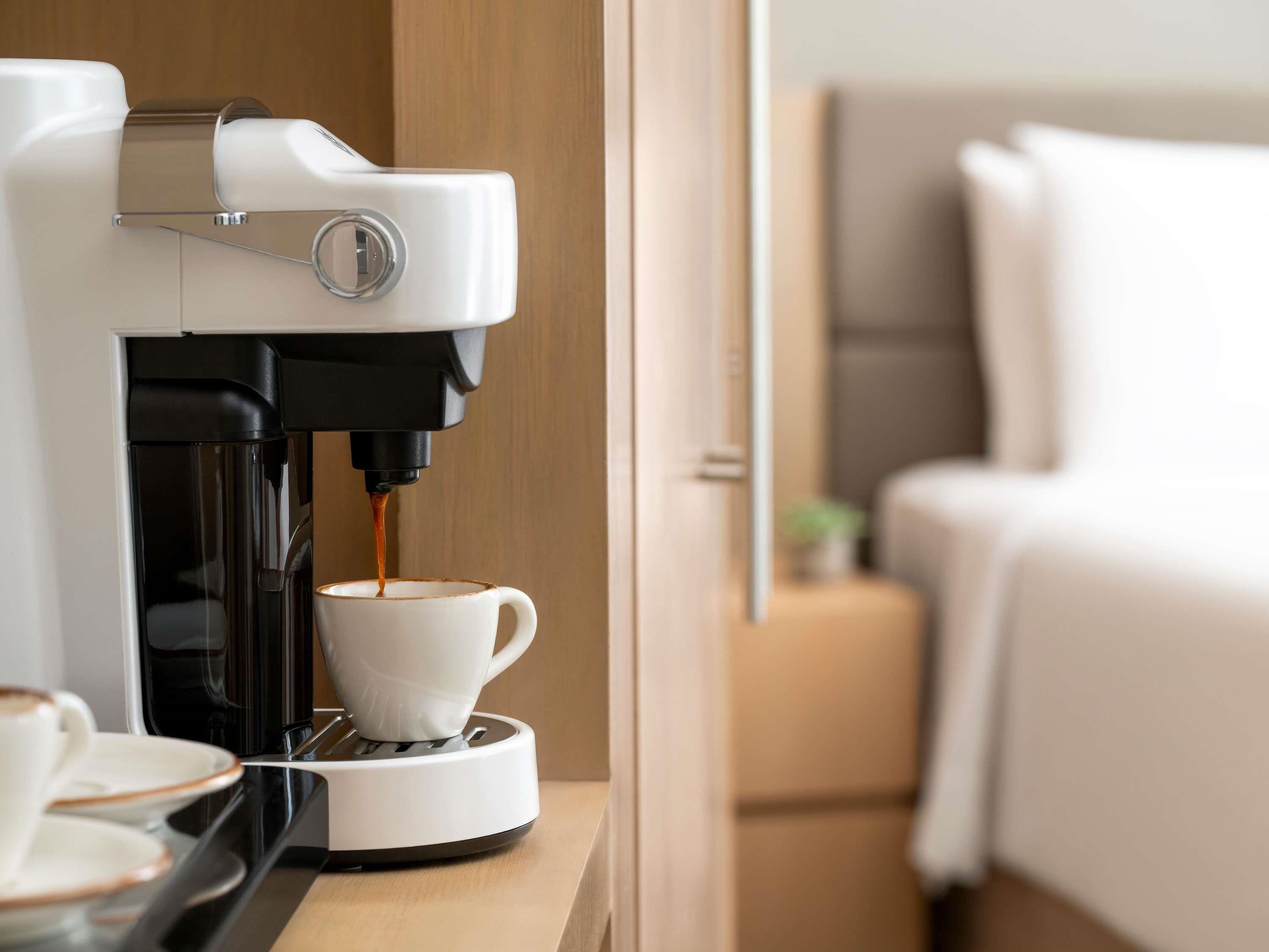 Uma máquina de café a servir café numa chávena, com uma cama e uma mesa de cabeceira ao fundo.