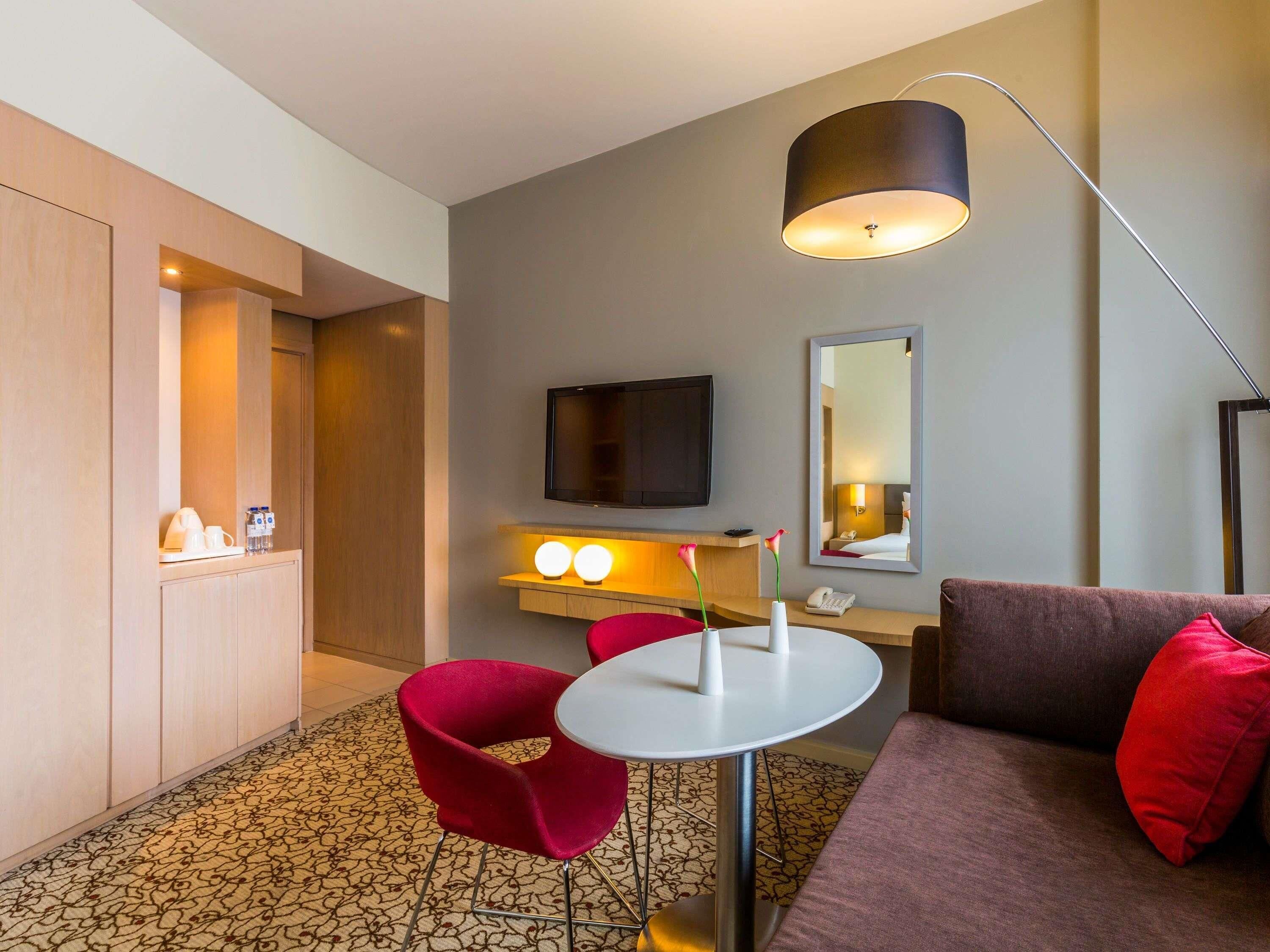 Een hotelkamer met een bank, een tafel met stoelen, een televisie, een spiegel en een lamp.