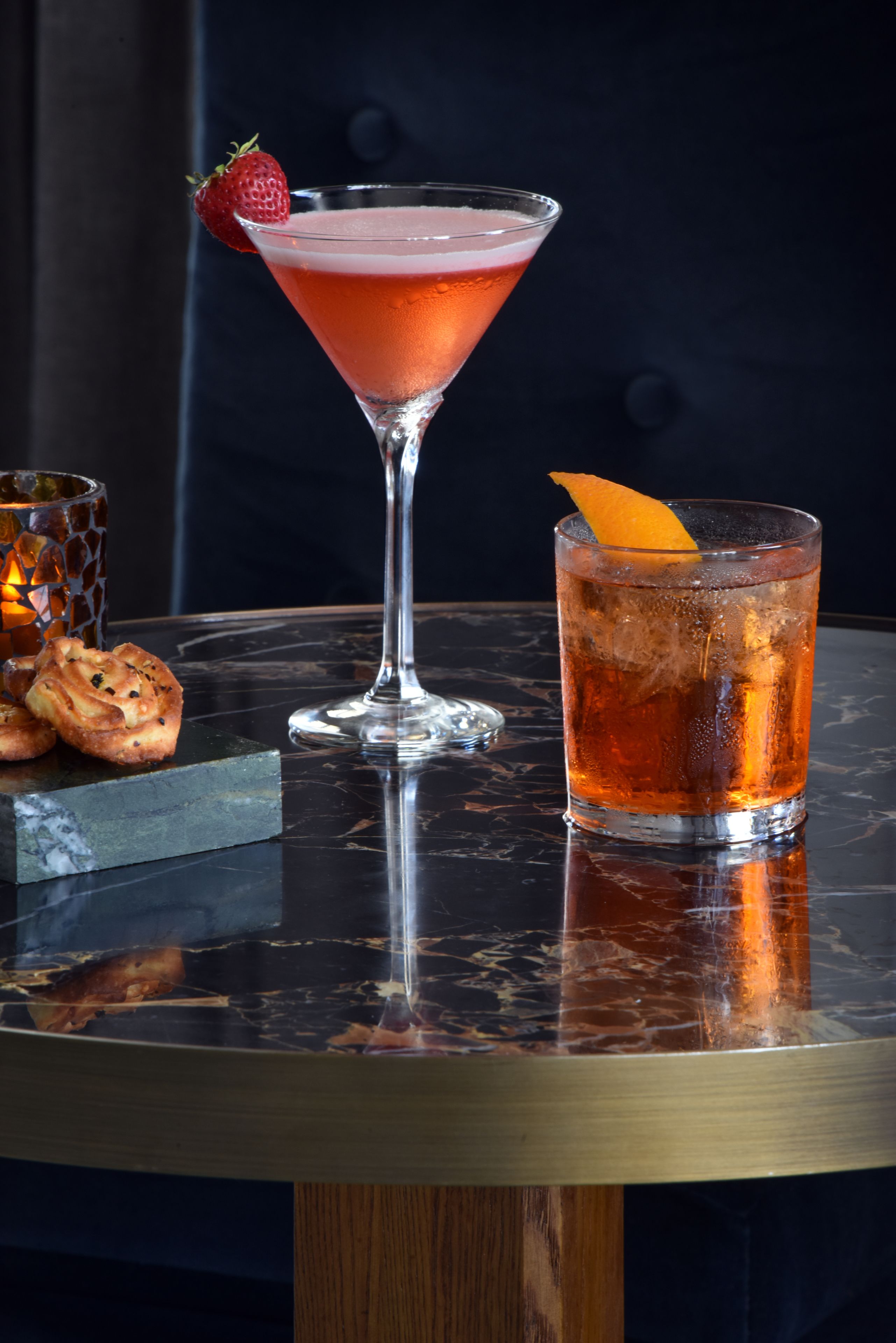 Una copa de martini adornada con fresas y una cáscara de naranja sobre una superficie reflectante.