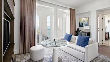 A modern living room with a round table, a sofa with blue cushions, a television, and a large window with curtains.