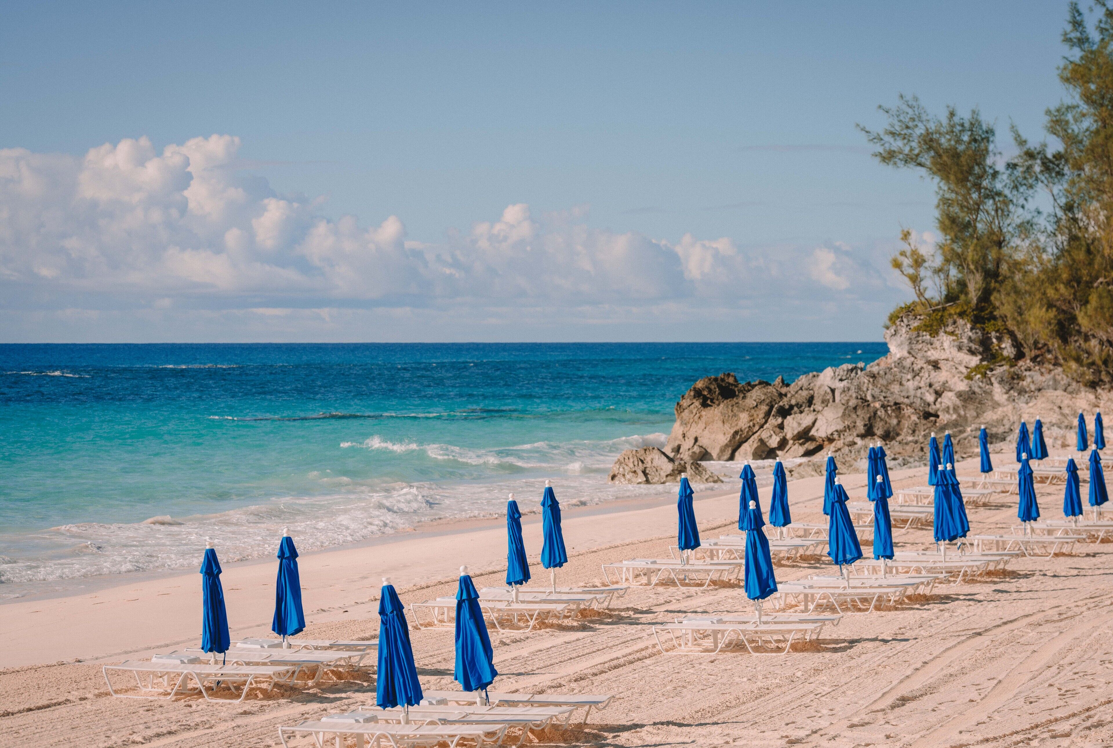 Ein Strand mit Reihen aus weißen Liegestühlen und blauen Sonnenschirmen, einer felsigen Klippe und dem Meer.
