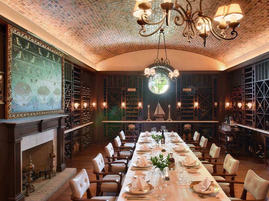 Una larga mesa de comedor dispuesta para una cena formal, rodeada de sillas, con una chimenea y estanterías para vinos al fondo.