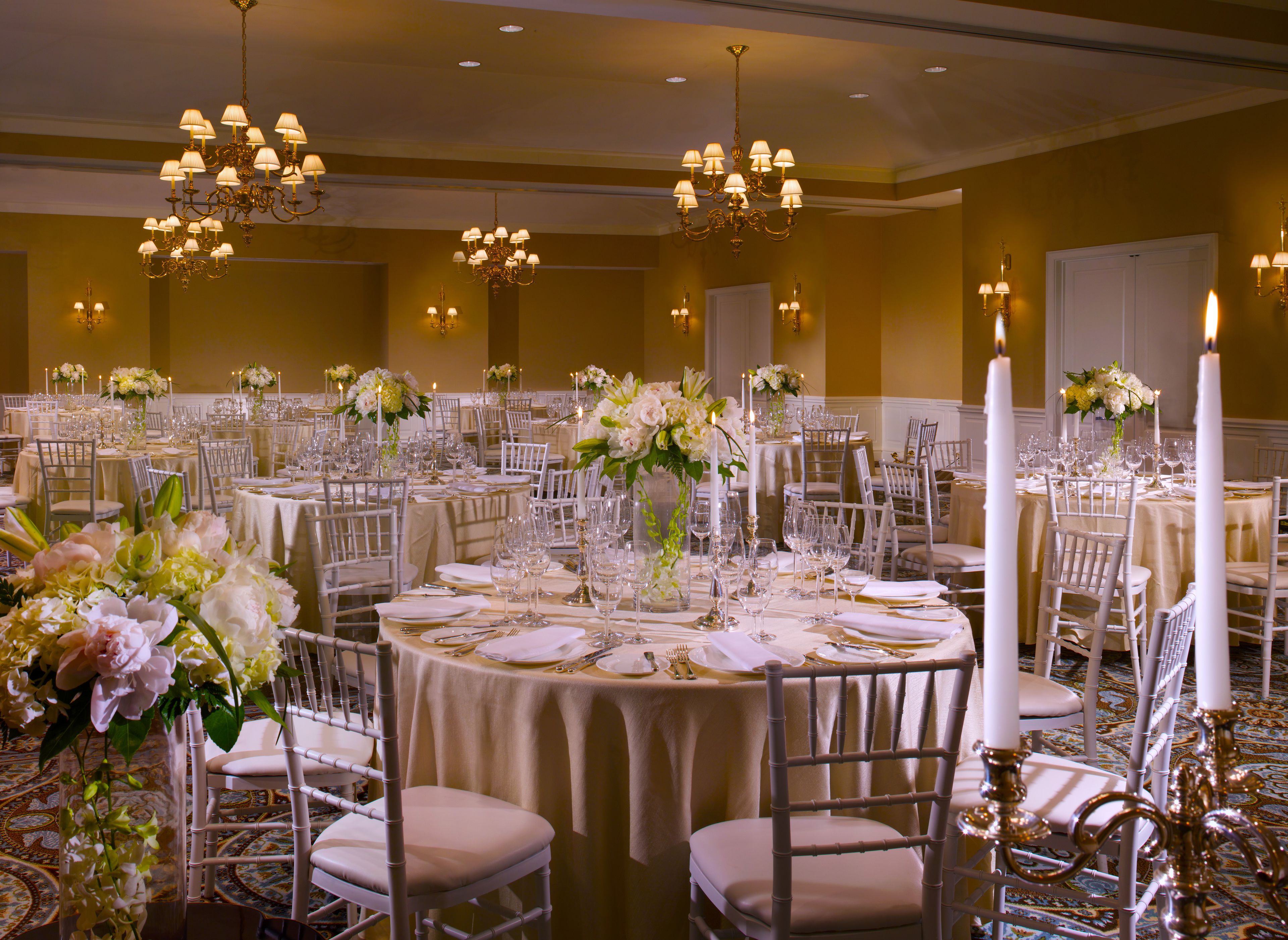 Un salón de banquetes con mesas redondas dispuestas para comer, sillas blancas, candelabros y centros de mesa con flores.