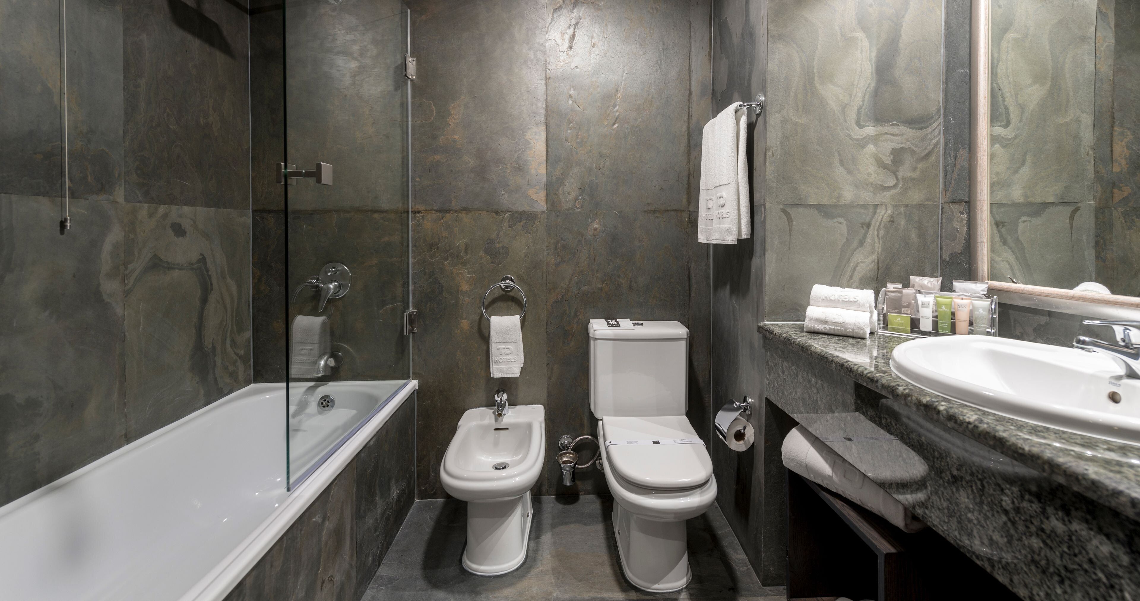 A modern bathroom with a bathtub, bidet, toilet, and granite countertop with a sink.