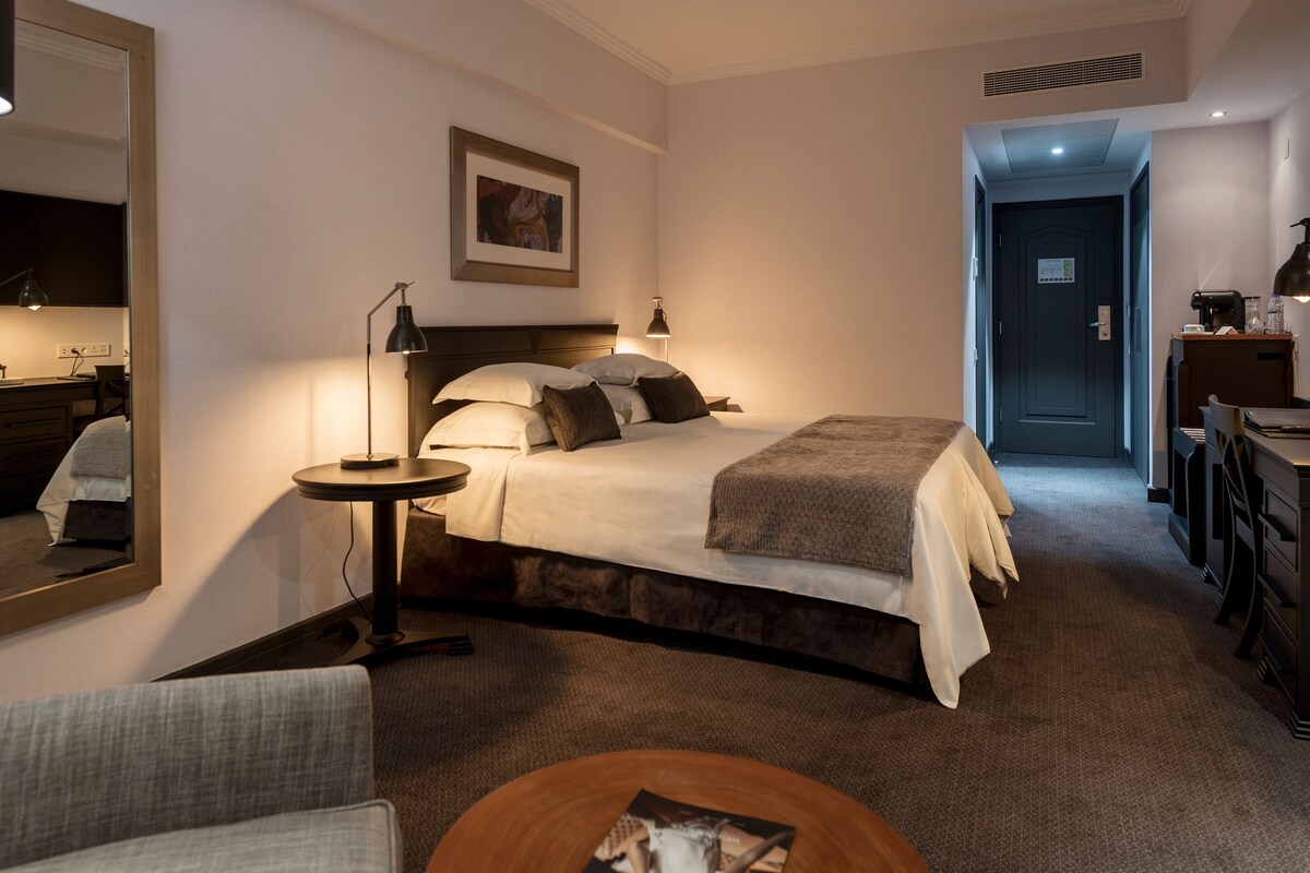 A hotel room with a bed, a desk, a chair, a round coffee table, and a mirror.