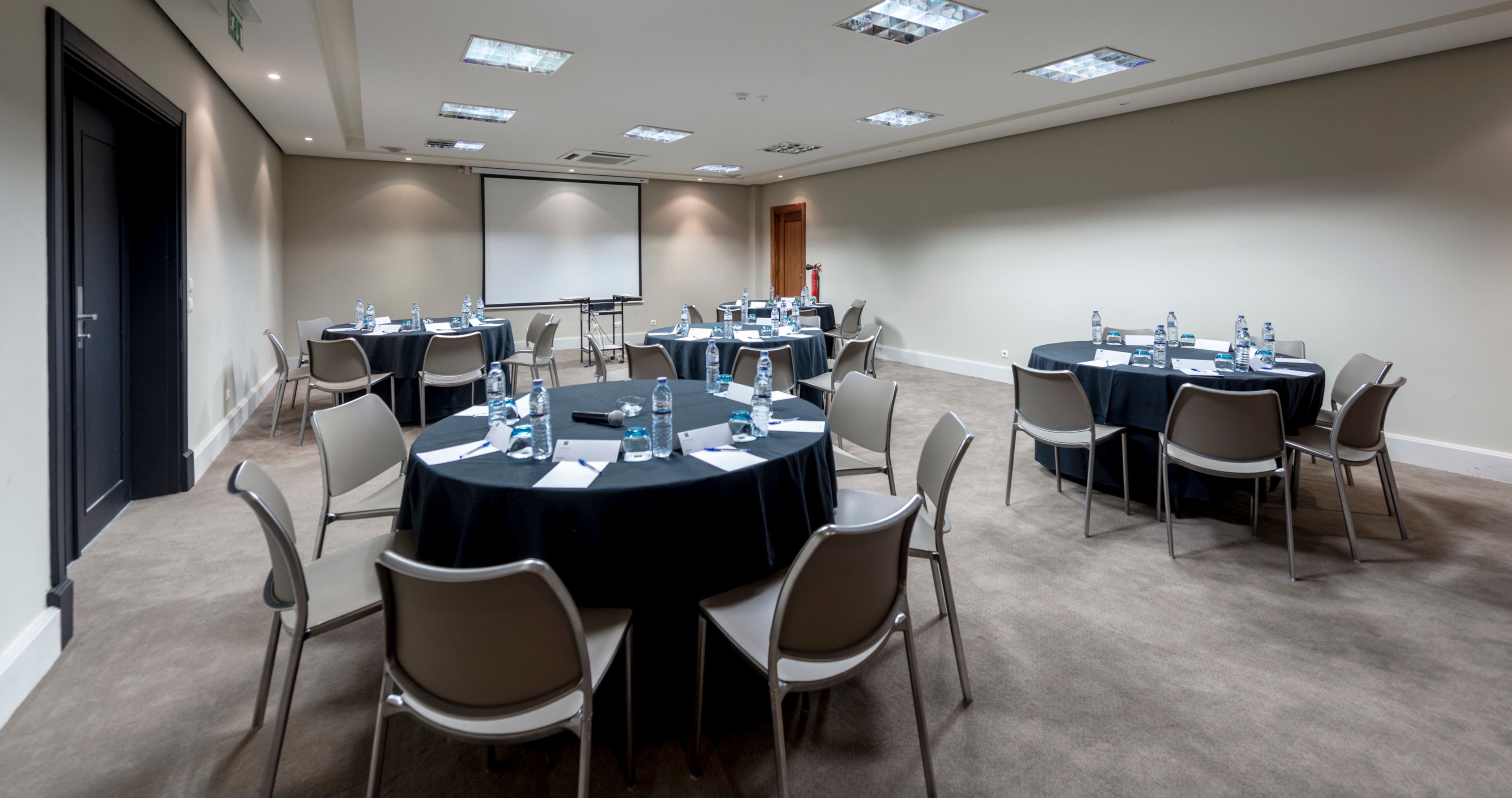 Un salón de conferencias con mesas redondas, sillas y una pantalla para proyector.