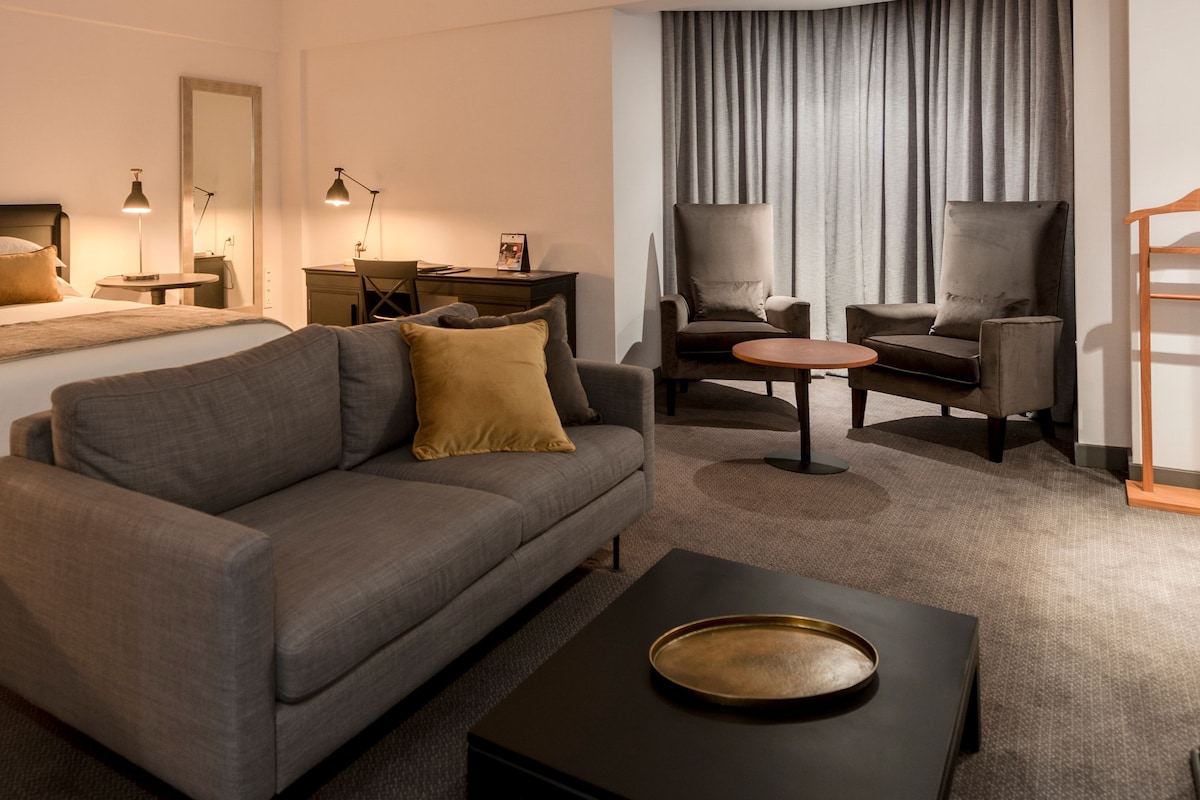 A modern hotel room with a sofa, two armchairs, a small round table, a bed, a desk, and a mirror.