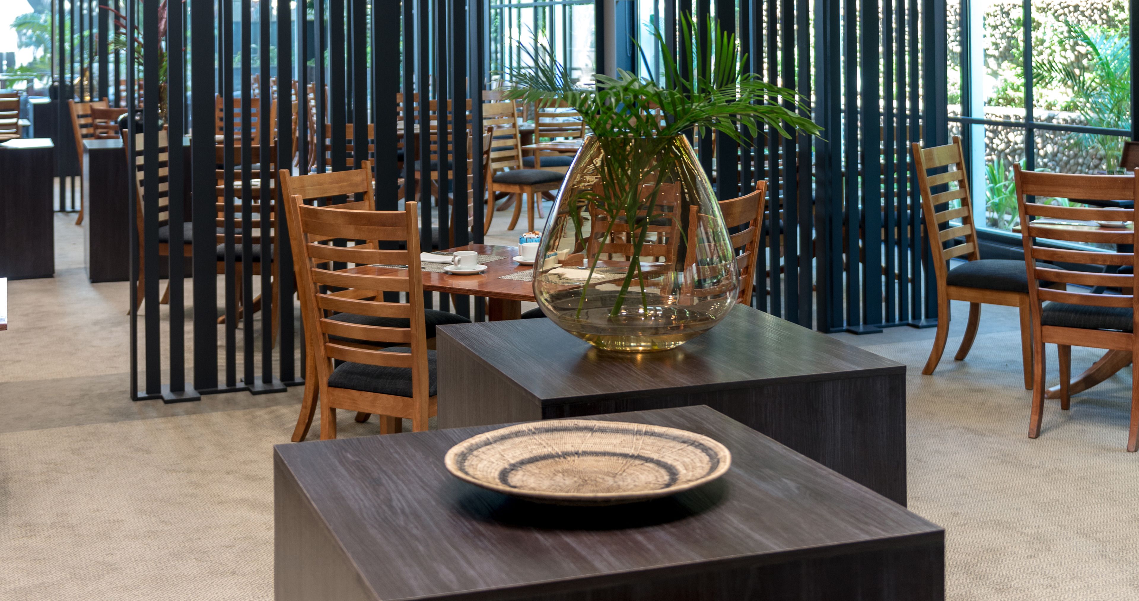 Un comedor con sillas y mesas de madera, un jarrón decorativo y un plato tejido sobre una mesa.