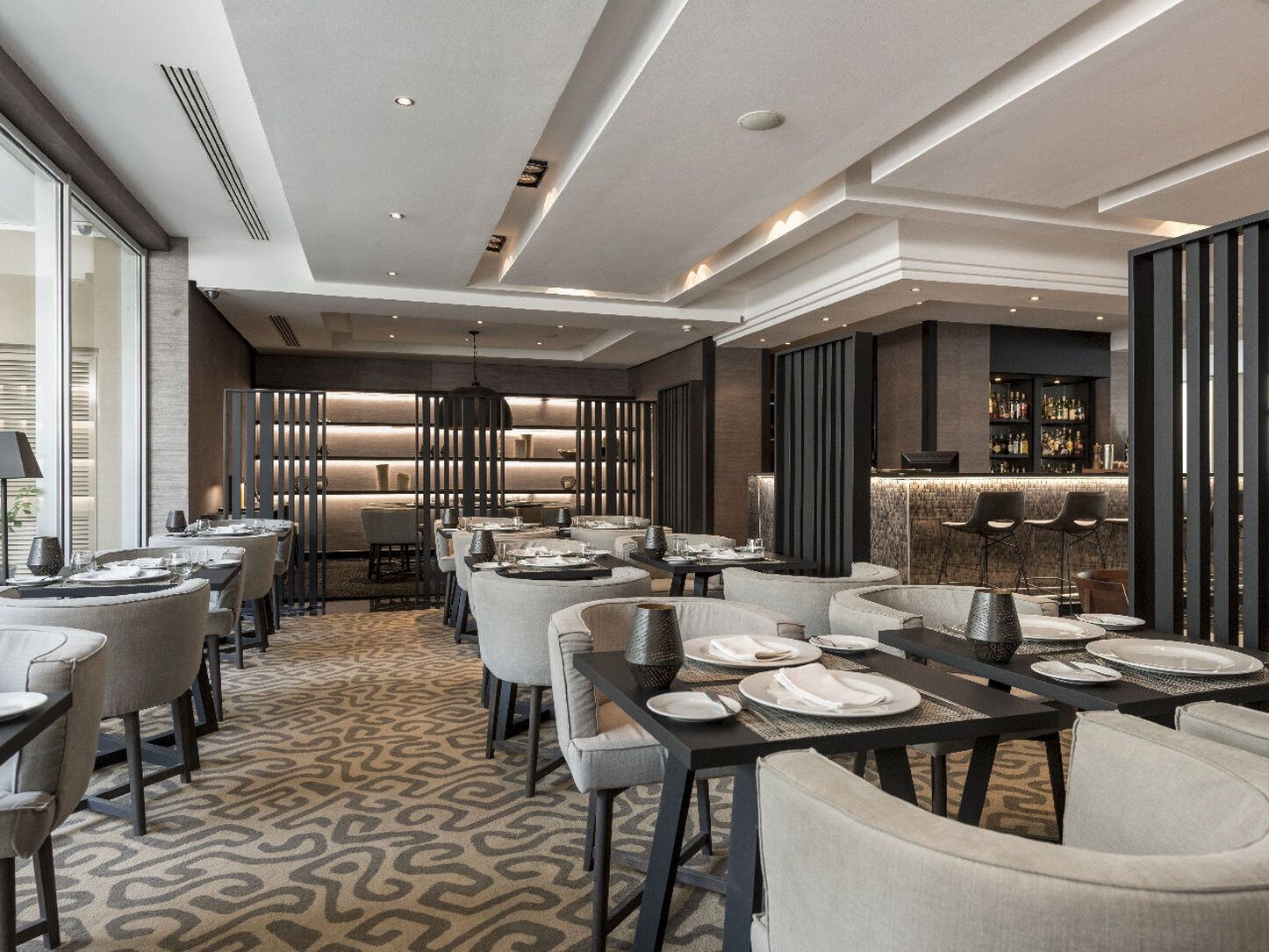 Een modern restaurant met een bar, voorzien van een patroon vloerkleed, eigentijds meubilair en een goed verlicht interieur.