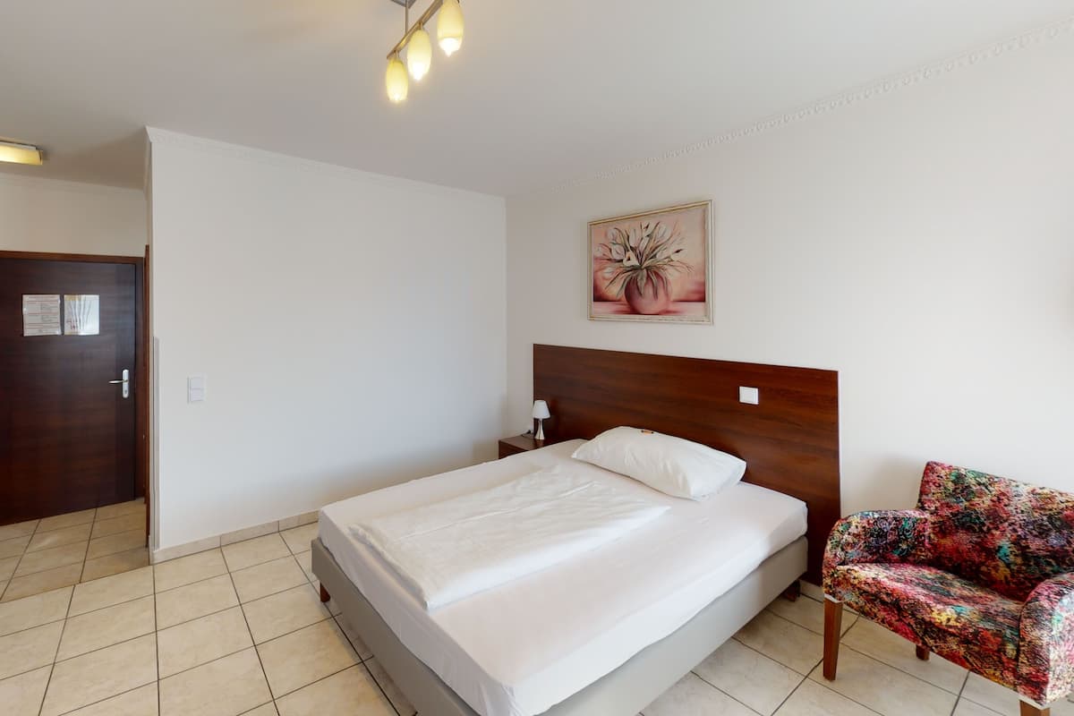 A hotel room with a bed, a nightstand, a chair, a door, and a painting on the wall.