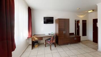 A hotel room with a wooden desk, a chair, a wardrobe, a television, and a trash bin.