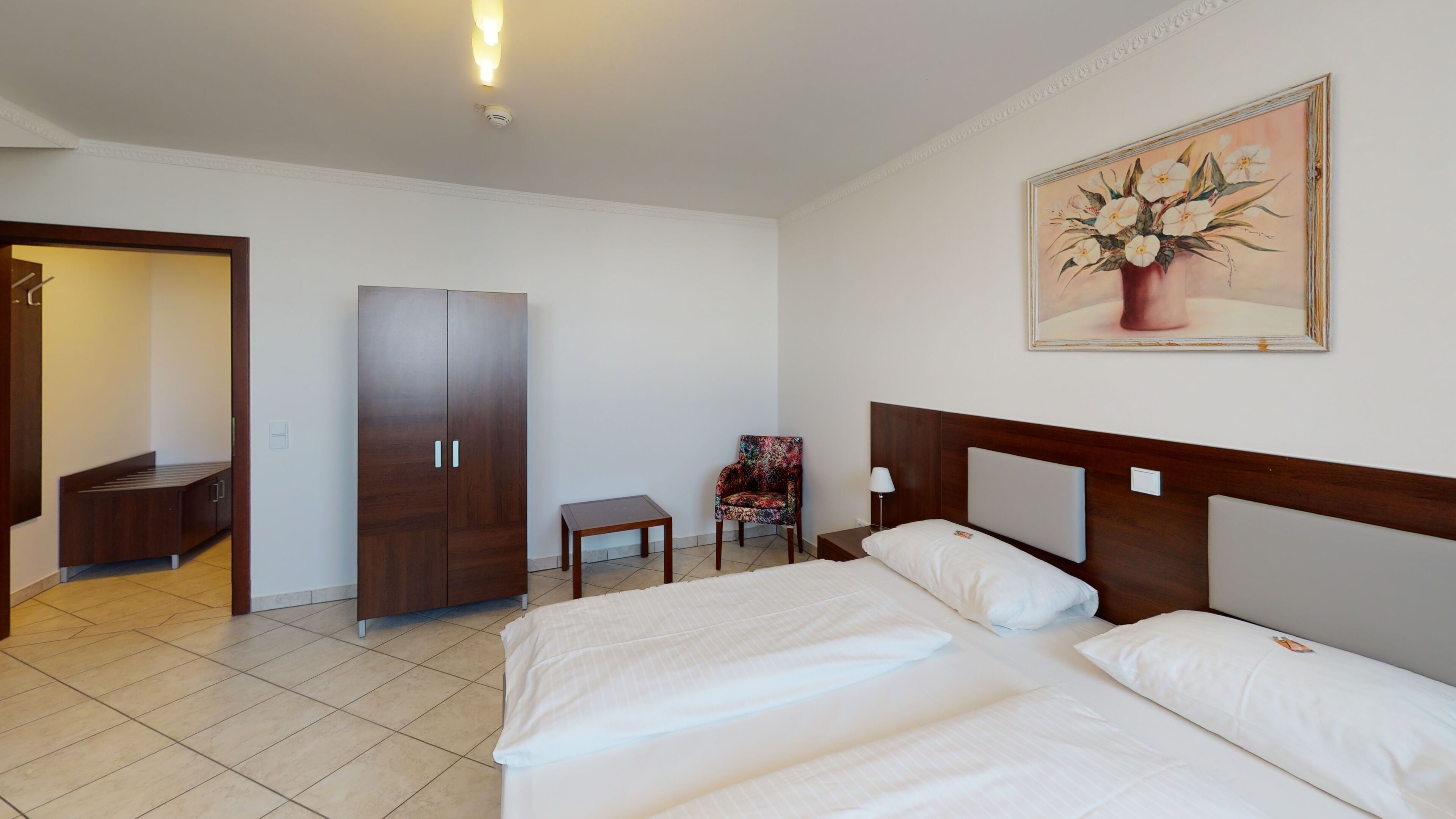 Una habitación de hotel con una cama, un armario, una mesita, una silla y un cuadro en la pared.