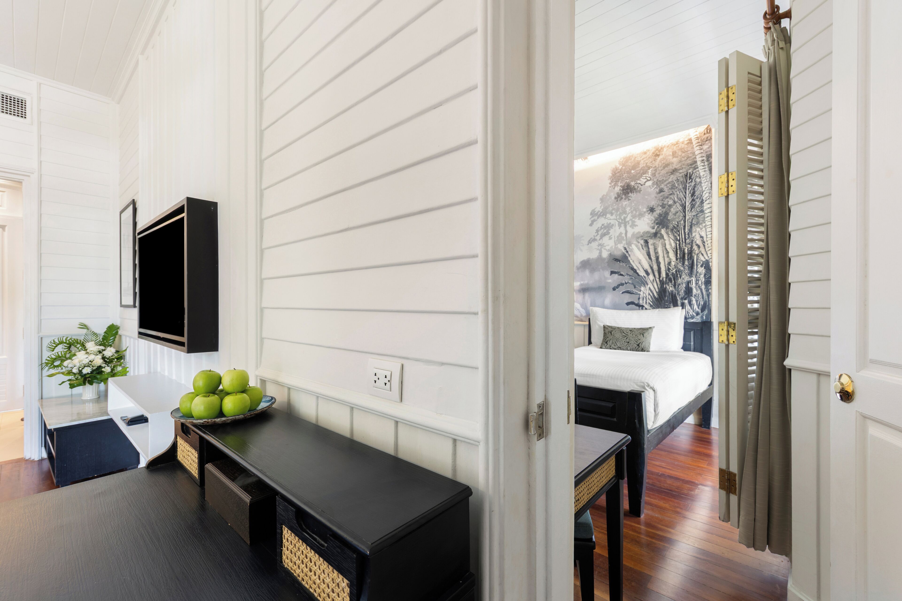 Una habitación con una cama, un escritorio con computadora, un televisor en la pared y una ventana con vista a árboles.