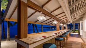 Un bar en la playa con taburetes de madera y una barra larga, con vistas al océano.
