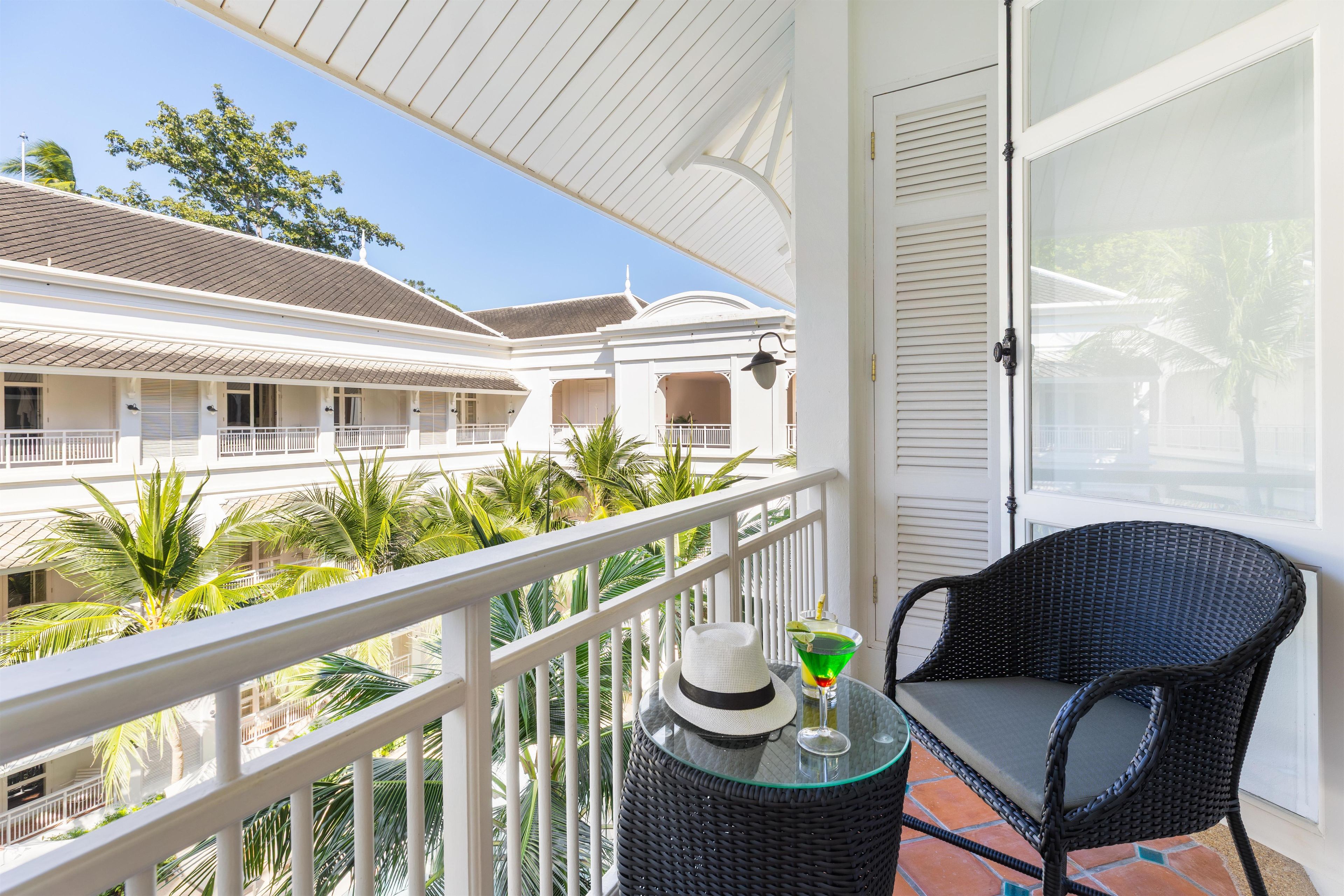 A balcony with a wicker chair, a glass table with a hat and a drink, and a view of a building with balconies and palm trees.