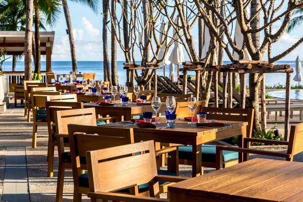Un restaurante frente a la playa con mesas y sillas de madera, dispuesto para comer con vistas al océano.