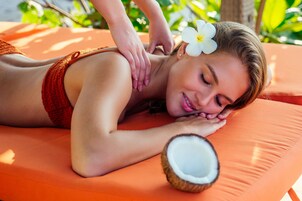 A person lying on an orange massage chair with a coconut and a white flower on their head.