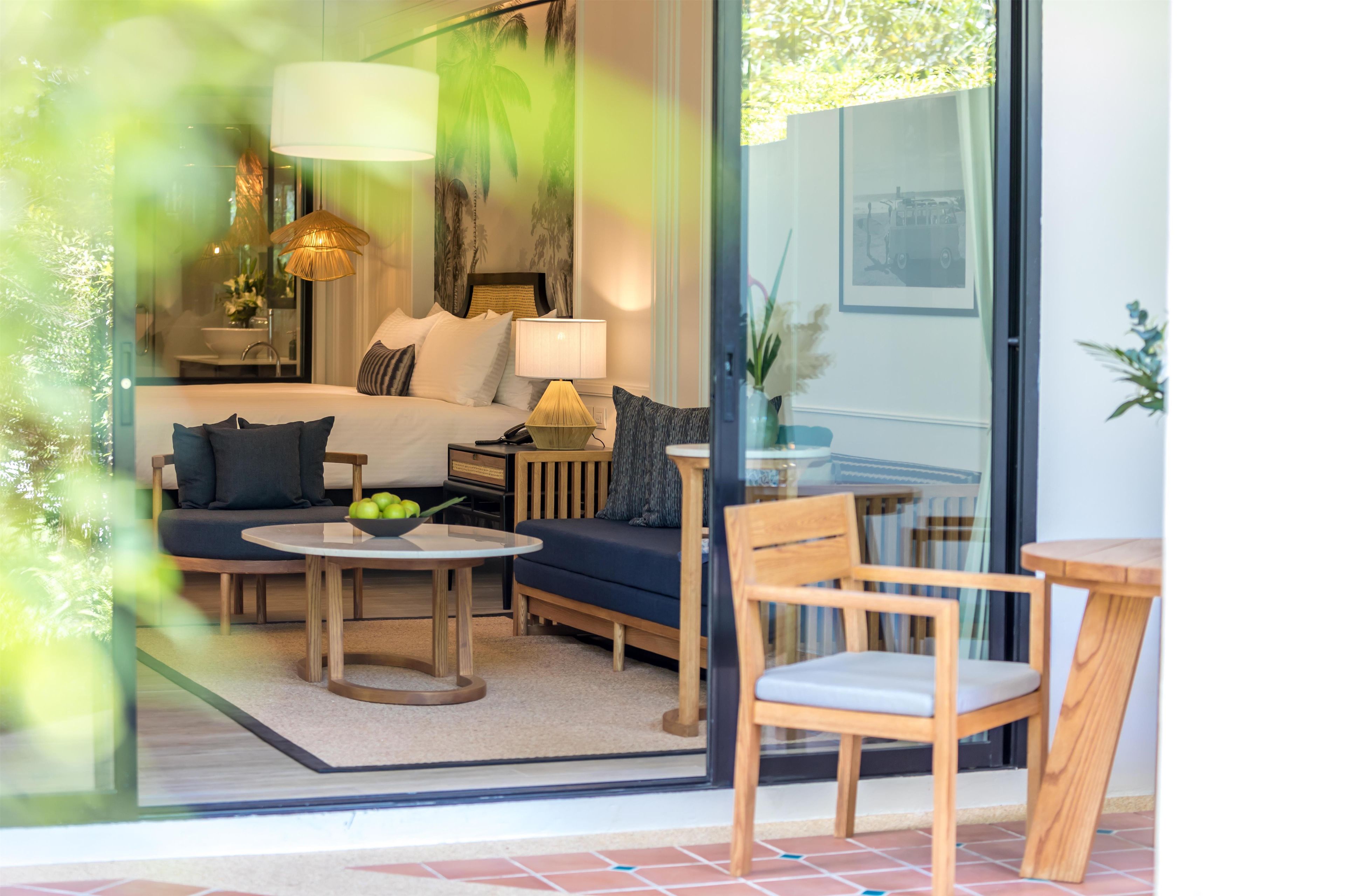 Une chambre d’hôtel moderne, équipée d’un lit, d’un canapé, d’une table basse et d’un fauteuil. Une fenêtre offre une vue sur les palmiers.