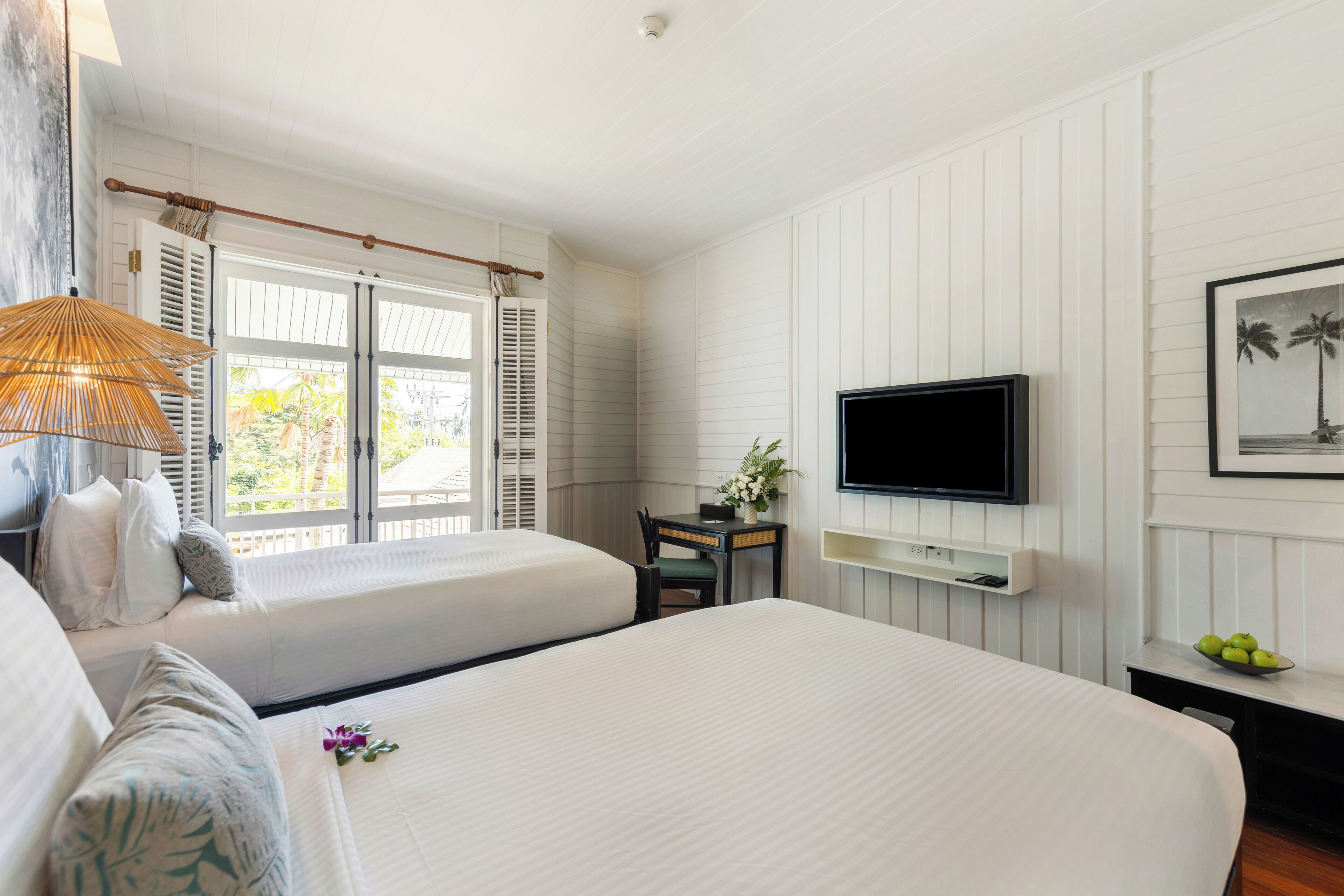 A hotel room with two beds, a TV, a desk, and a window with shutters.