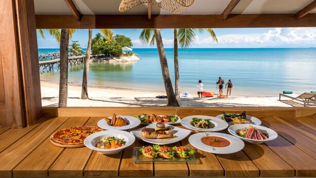 Una mesa de madera con varios platos y vista a una playa con palmeras y un muelle.