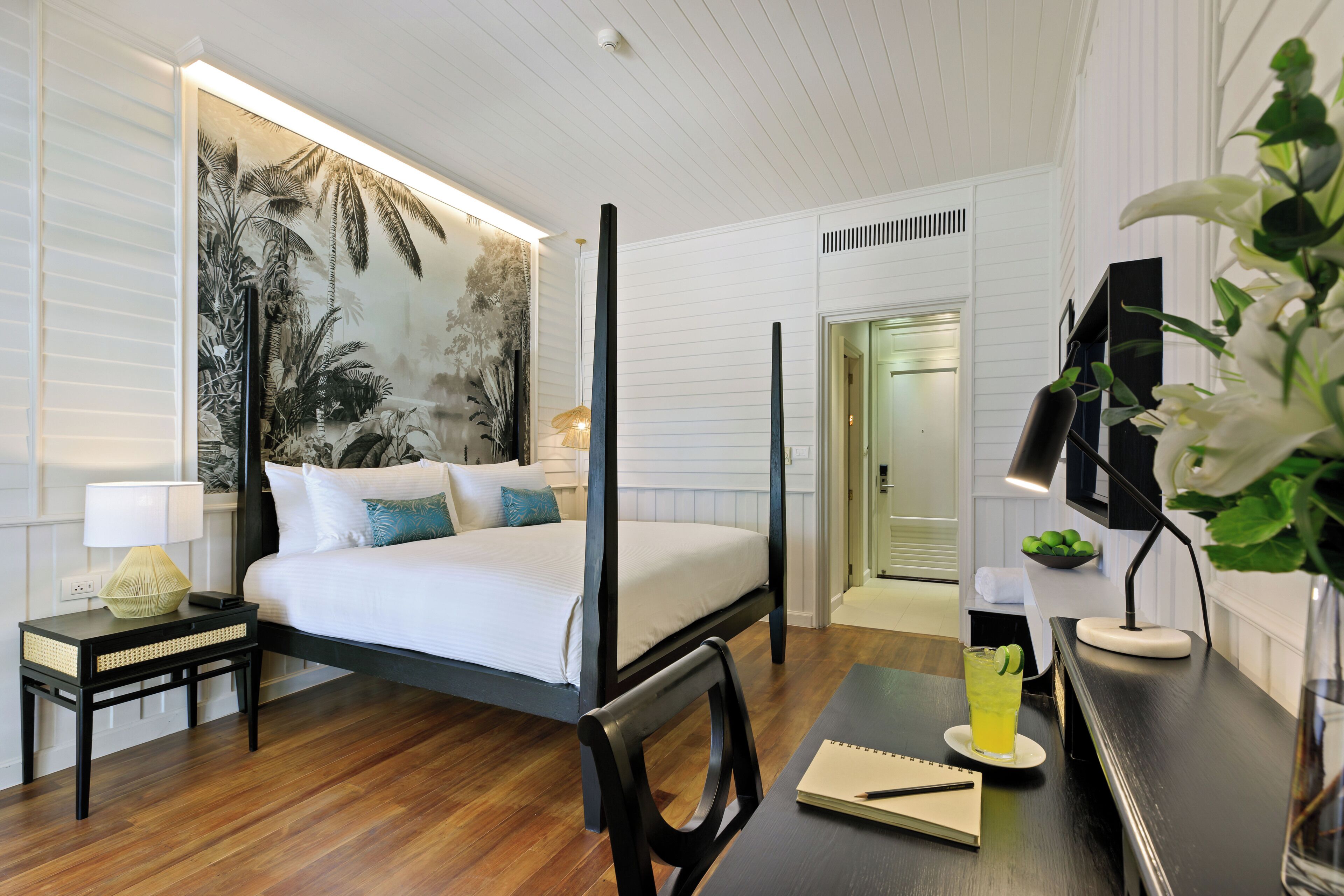 A four-poster bed, a desk with a notebook, a glass of drink, and a wall with a tropical mural.