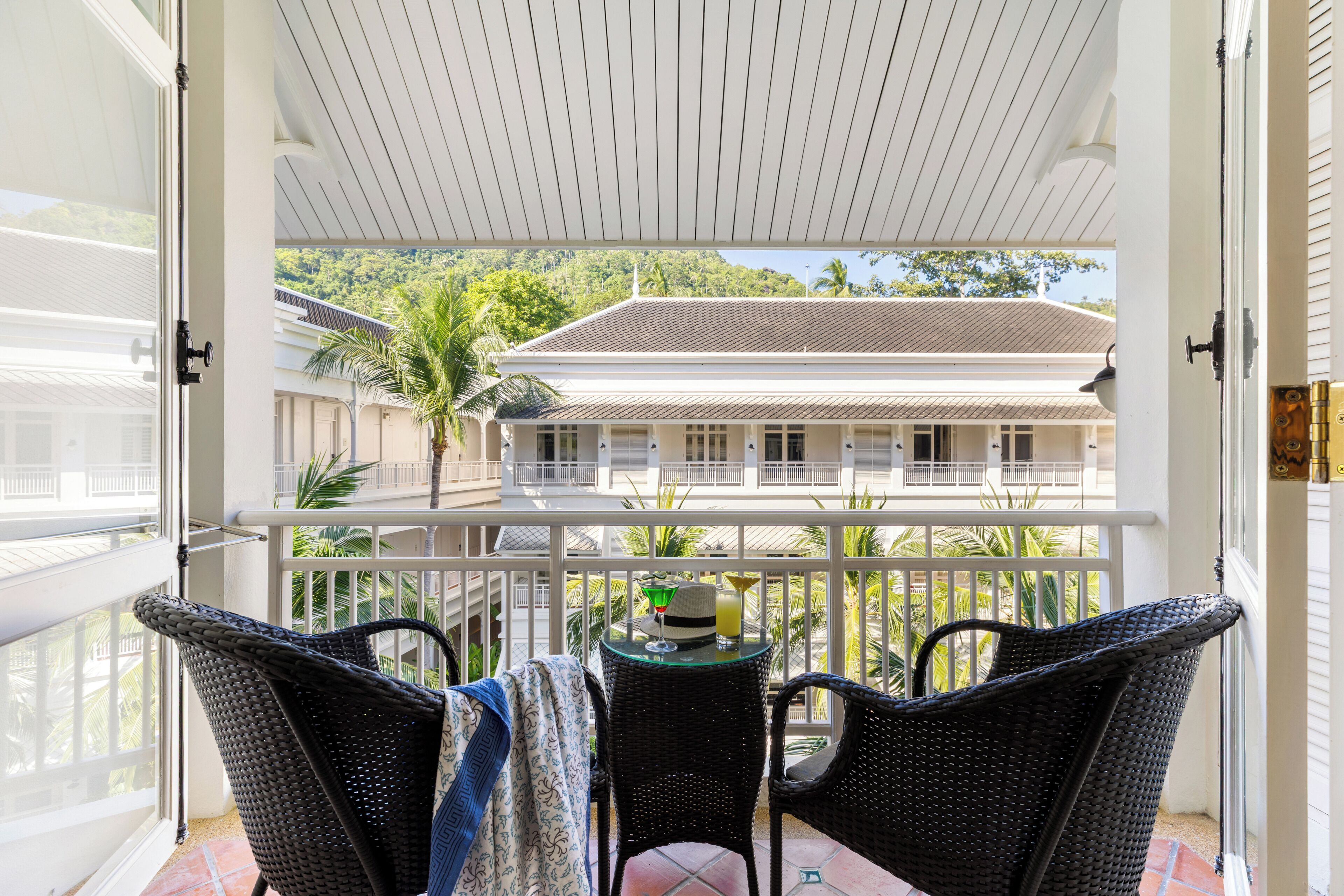 A balcony with wicker chairs, a small table with a drink, and a view of a building with a balcony.