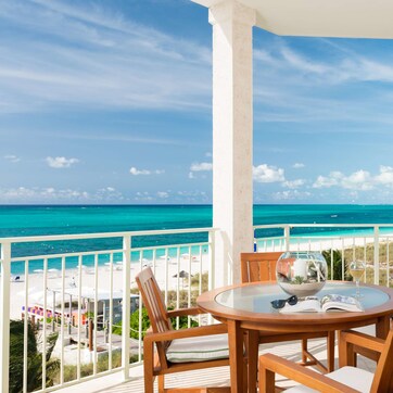 A round wooden table with four chairs, a glass bowl, a wine glass, and an open book on a balcony overlooking a beach and ocean.