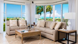A spacious living room with a large window offering a view of the ocean and palm trees. There is a beige sofa with decorative pillows, a wooden coffee table, and a small side table with a lamp.
