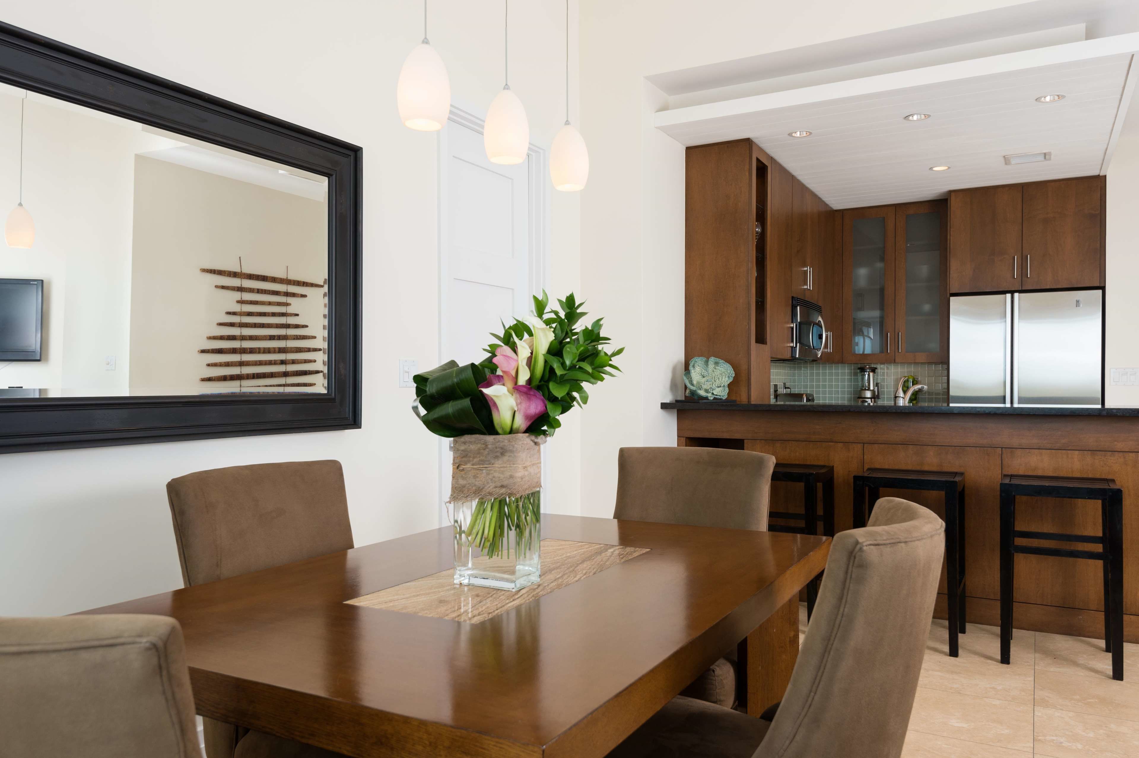 Un comedor con una mesa de madera, sillas y un jarrón con flores. Se ve una cocina al fondo con armarios de madera y un refrigerador.
