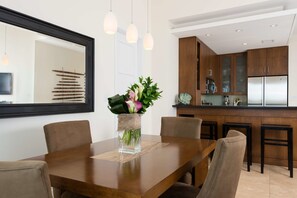 A dining area with a wooden table, chairs, and a vase with flowers. There is a kitchen in the background with wooden cabinets and a refrigerator.