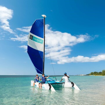 A sailboat with a blue and white sail on a clear day, with calm turquoise waters and a beach in the background.