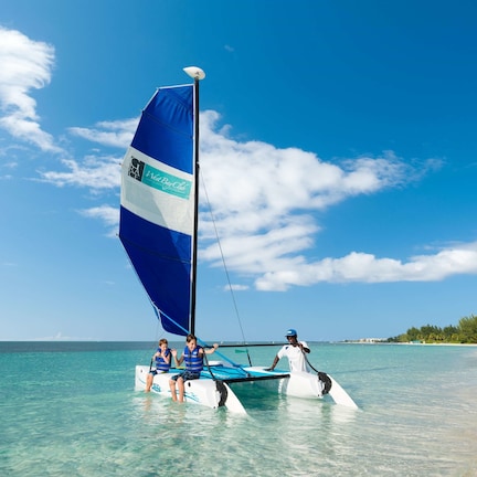 A sailboat with a blue and white sail on a clear day, with calm turquoise waters and a beach in the background.