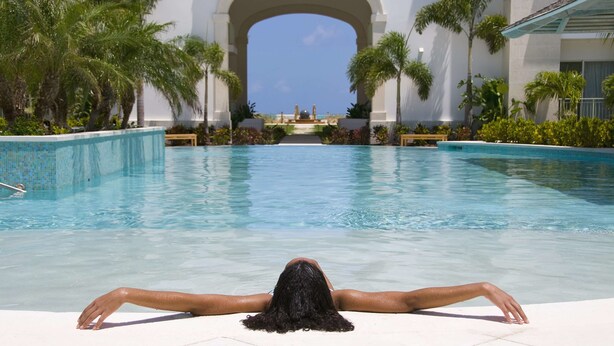 A pool area with a clear view of the ocean through an archway, surrounded by palm trees and a tiled deck.