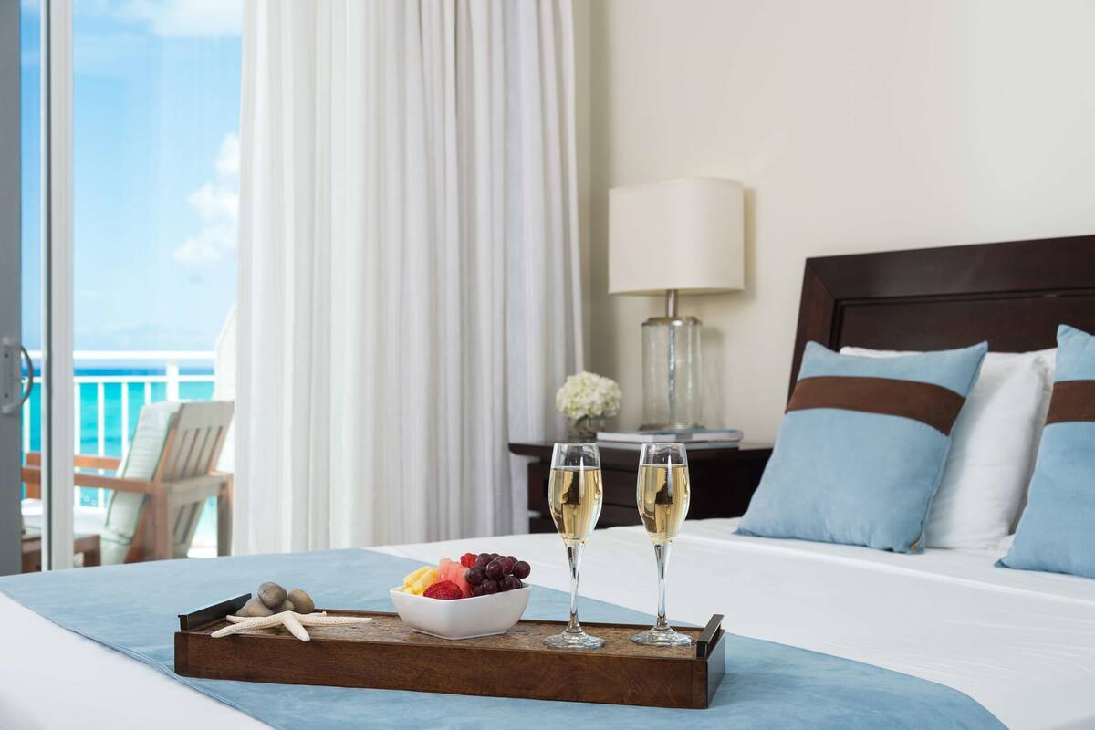 A hotel room with a bed, a tray with fruit and two glasses of champagne, a nightstand with a lamp, and a view of the ocean through large windows.