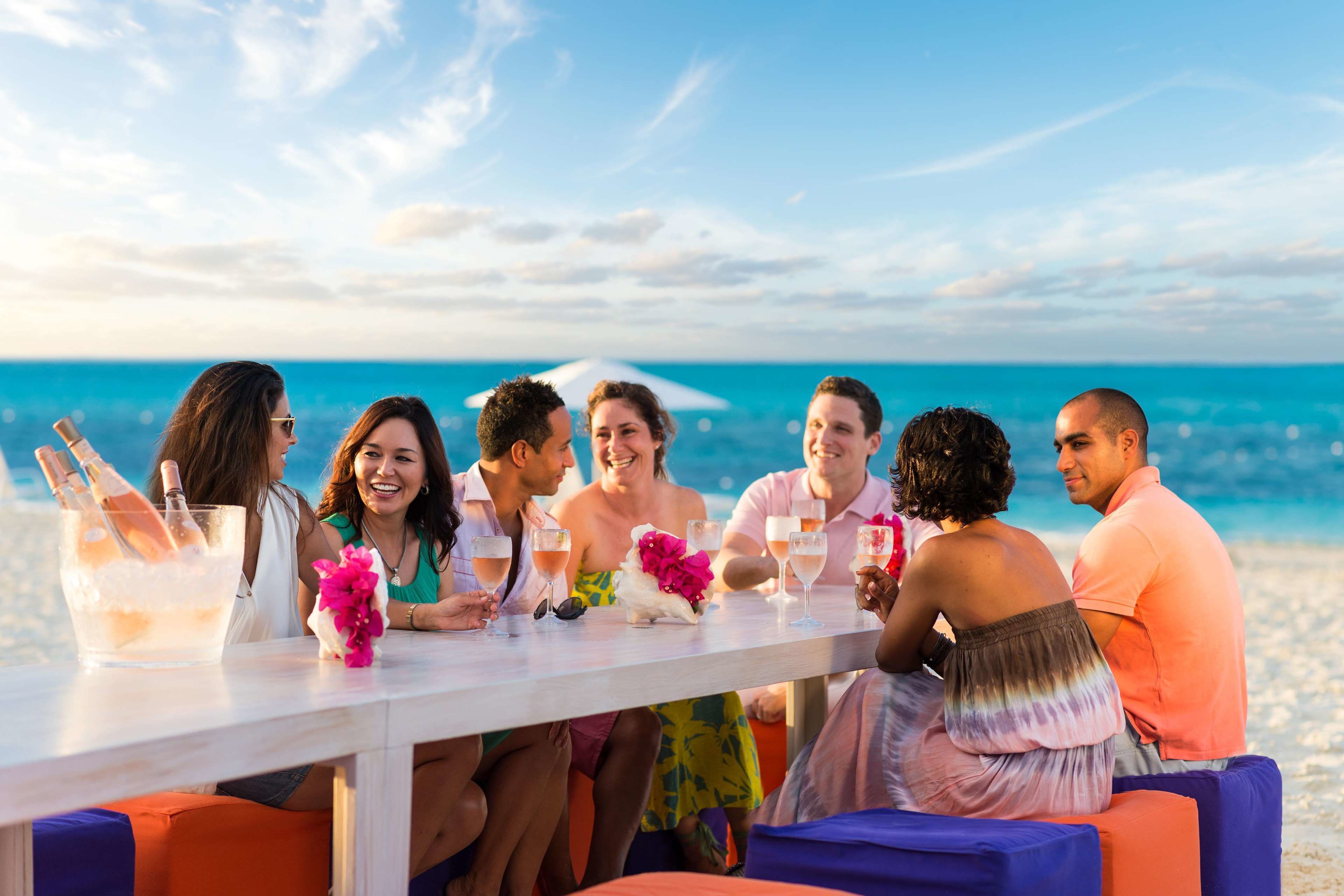 A group of people seated at a table on a beach with drinks and a flower arrangement.