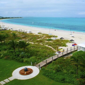 A beach resort with a wooden boardwalk leading to a white building, surrounded by lush greenery and a clear blue sea.