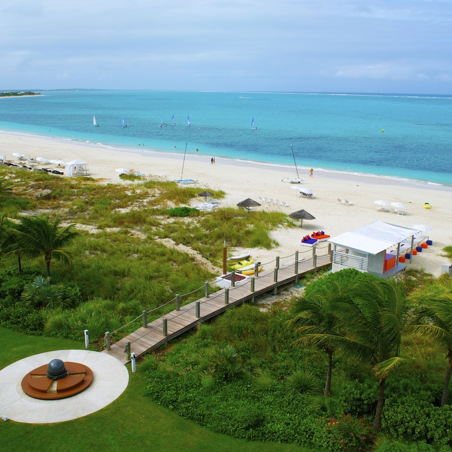 A beach resort with a wooden boardwalk leading to a white building, surrounded by lush greenery and a clear blue sea.