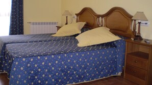 Una cama doble con colcha estampada, dos lámparas de noche, una mesita de noche con lámpara y una pared con dos cuadros enmarcados.