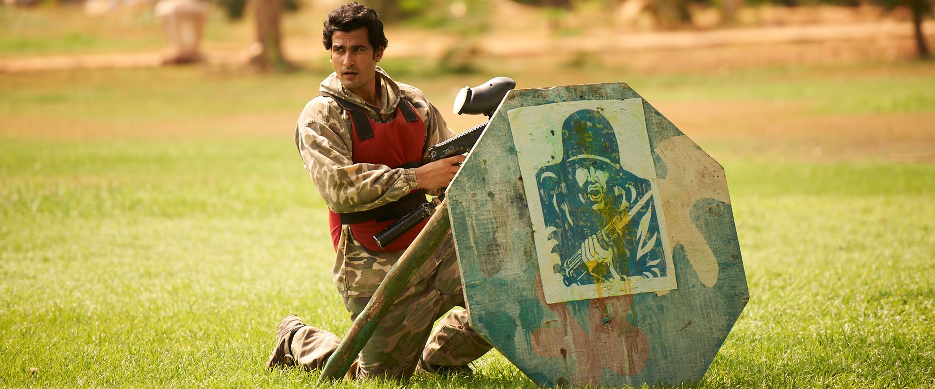 A person in camouflage attire kneeling on grass, holding a rifle.