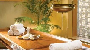 Een houten massagetafel met handdoeken en een massageapparaat, een potplant en een raam met een lattenroede.