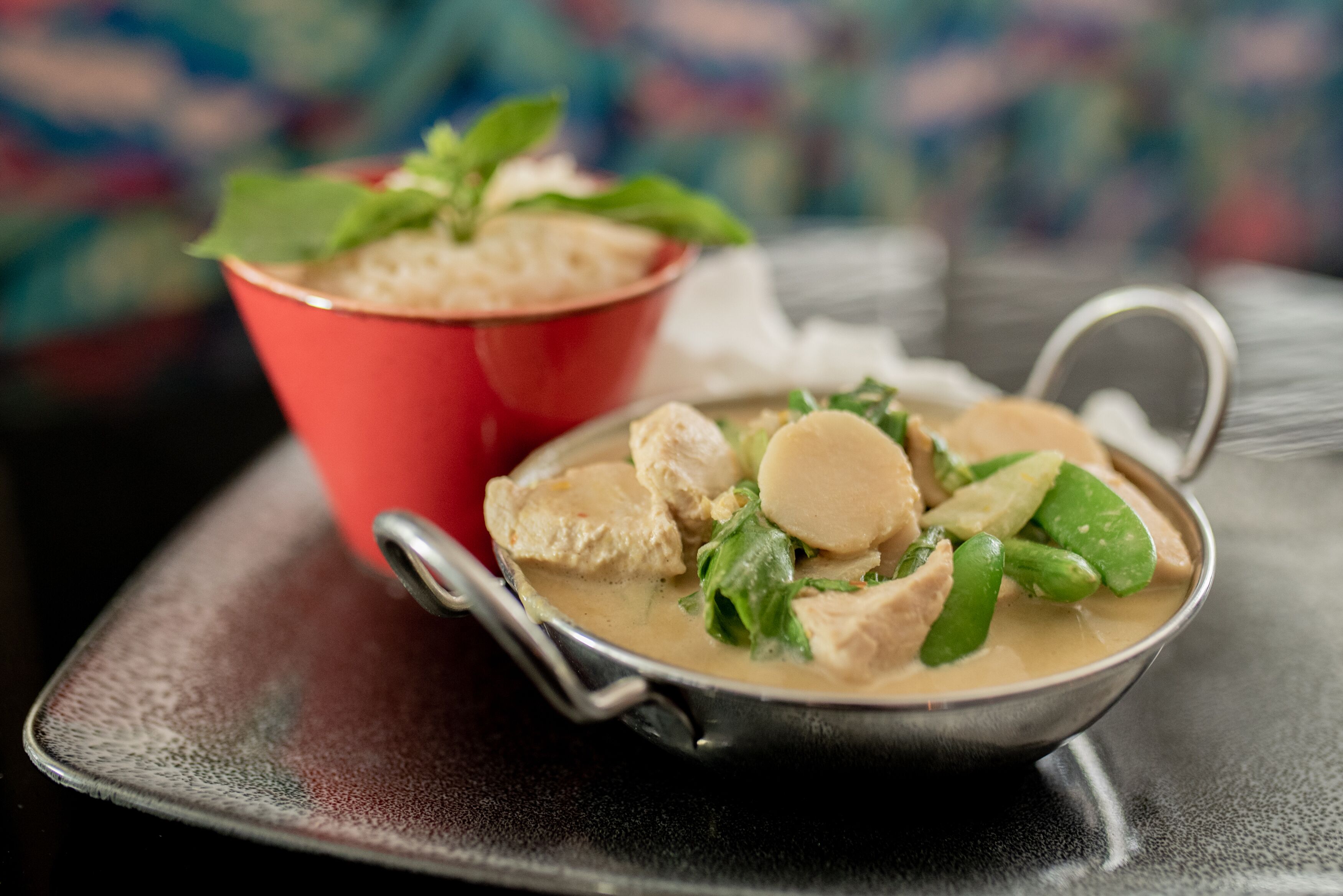 A bowl of soup with chunks of meat and vegetables, a red cup, and a spoon on a table.
