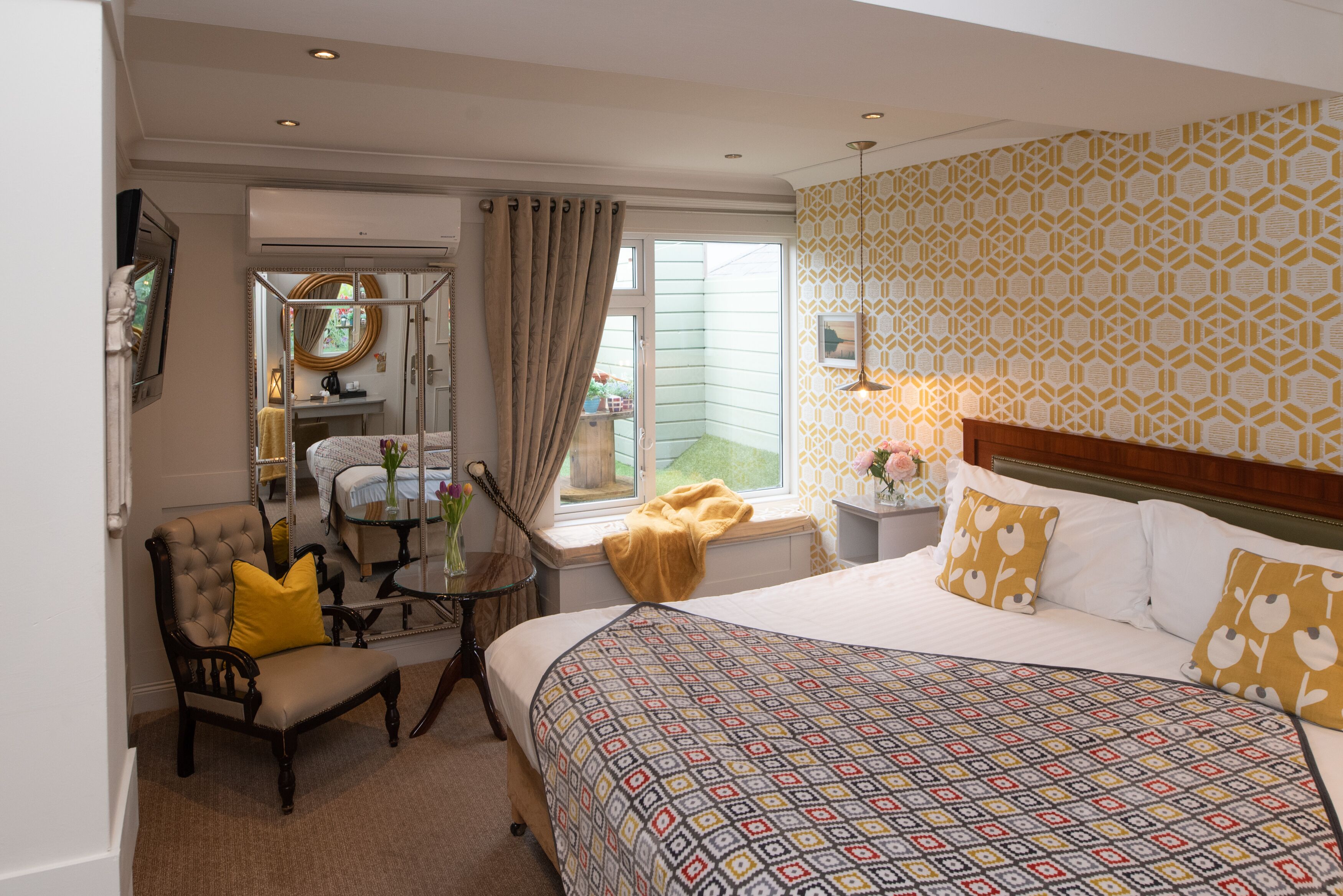 Een hotelkamer met een groot bed, een stoel met een geel kussen, een klein tafeltje met een vaas en een raam met uitzicht.